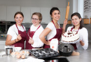 4 Frauen mit Küchenschürzen stehen nebeneinander, eine hält ein Nudelholz, eine hält eine Torte